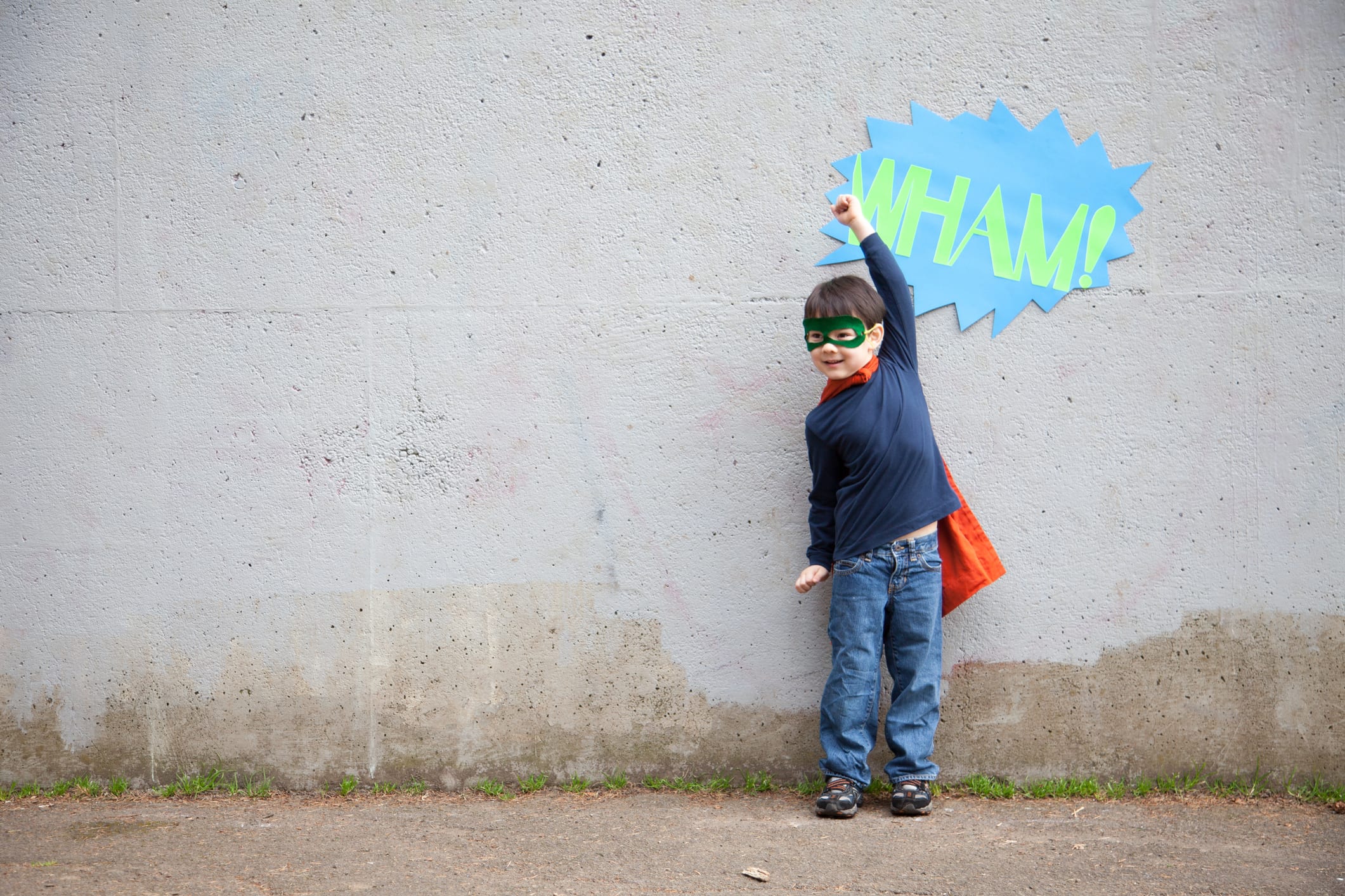 Young boy, with cape and mask, arm up, pretends to be a superhero in front of a wall with the word Wham! on it.